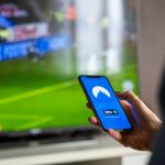 Pessoa segura um smartphone com um aplicativo de VPN aberto, enquanto uma transmissão esportiva aparece ao fundo em uma televisão.