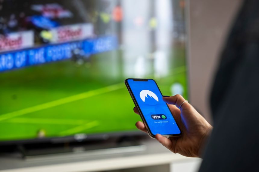 Pessoa segura um smartphone com um aplicativo de VPN aberto, enquanto uma transmissão esportiva aparece ao fundo em uma televisão.