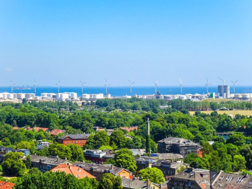 Vista aérea de turbinas eólicas próximas a uma cidade litorânea.