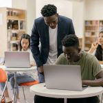 Estudantes em uma sala de aula interagem com uma professora que os orienta, enquanto laptops estão dispostos sobre as mesas.