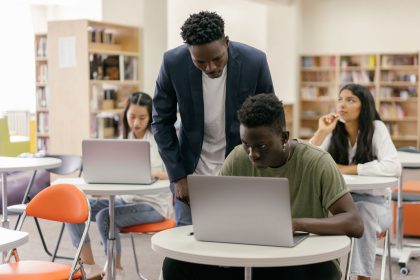 Estudantes em uma sala de aula interagem com uma professora que os orienta, enquanto laptops estão dispostos sobre as mesas.
