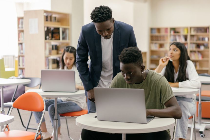 Estudantes em uma sala de aula interagem com uma professora que os orienta, enquanto laptops estão dispostos sobre as mesas.